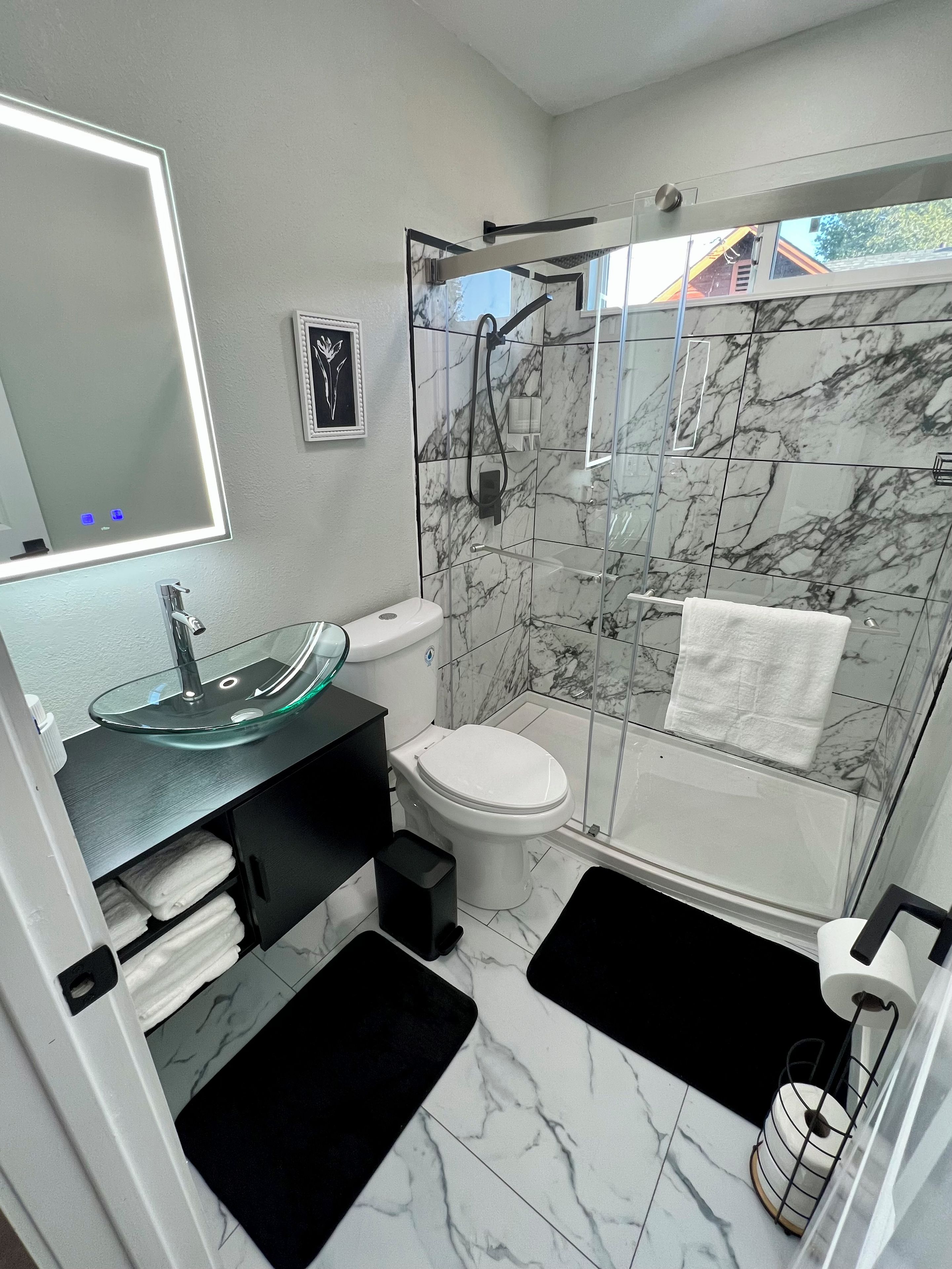 A bathroom with a glass shower enclosure, a marble-tiled floor, a toilet, a sink with a glass basin, and a towel rack.