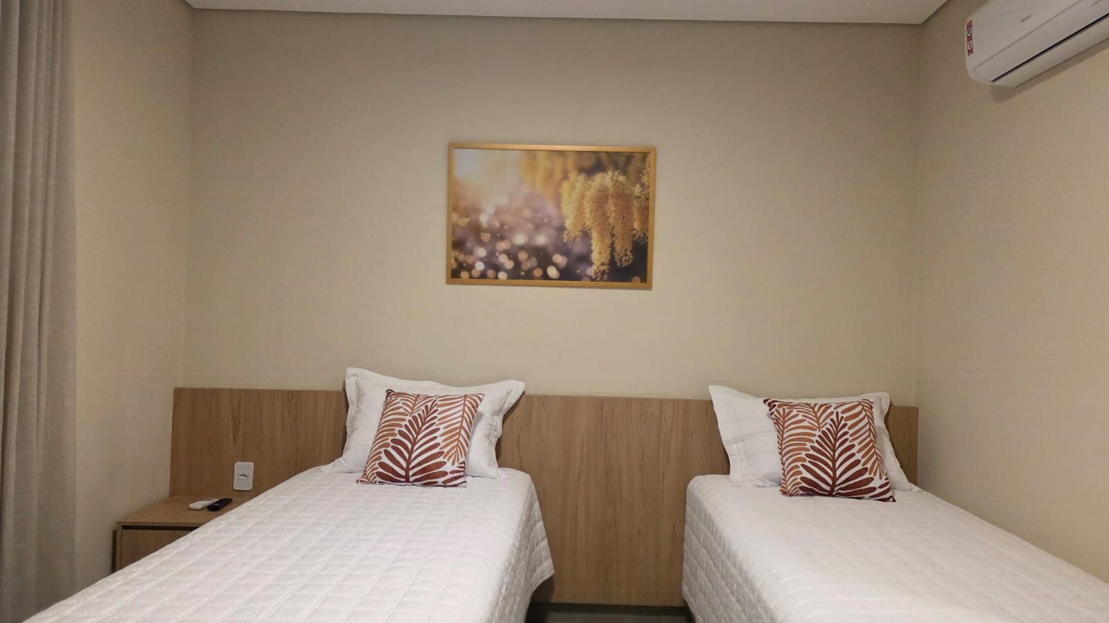 Habitación de hotel con dos camas, una mesita de noche, aire acondicionado montado en la pared y un cuadro sobre las camas.