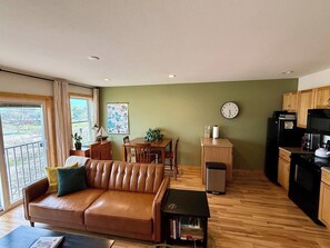 Ein Wohnzimmer mit einer Ledersofa, einem Esstisch mit Stühlen, einer Küchenzeile inklusive Schränken und Geräten sowie einem Blick durch eine Glastür.