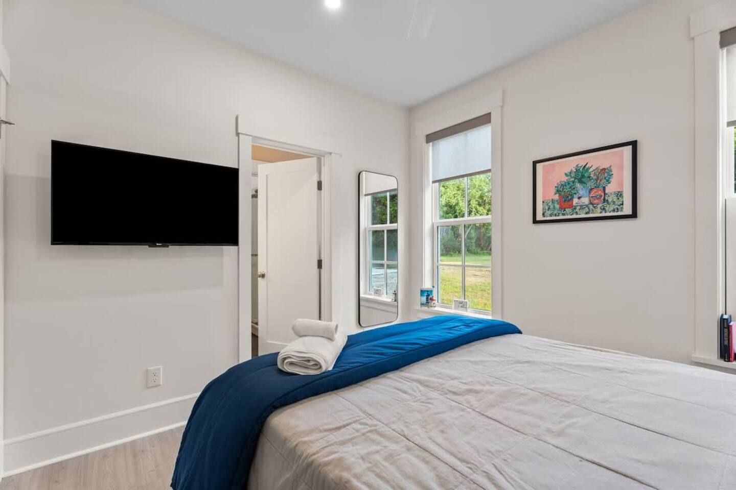 A bedroom with a bed, a television mounted on the wall, a door leading to another room, and a window with a view of greenery.