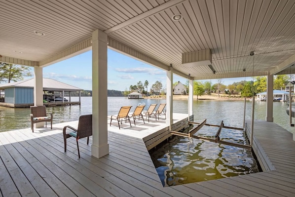 A covered dock with chairs and a small boat, overlooking a calm lake with houses in the background.