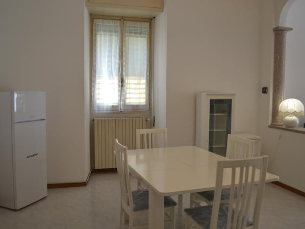 Une pièce avec une table et des chaises blanches, un radiateur, une fenêtre avec des rideaux et un meuble blanc.