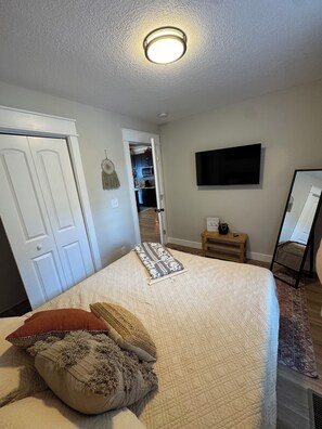 A bedroom with a bed, a flat-screen TV, a door, and a hallway leading to a kitchen. - 9139 Orange -2-bedroom w/ Loft - Melton Hill Lake & Marina -Close to downtown UT (Oak Ridge)