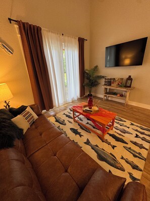 A living room with a brown leather sofa, a fish-patterned rug, a wooden coffee table, a flat-screen TV, and a window with curtains. - 9139 Orange -2-bedroom w/ Loft - Melton Hill Lake & Marina -Close to downtown UT (Oak Ridge)