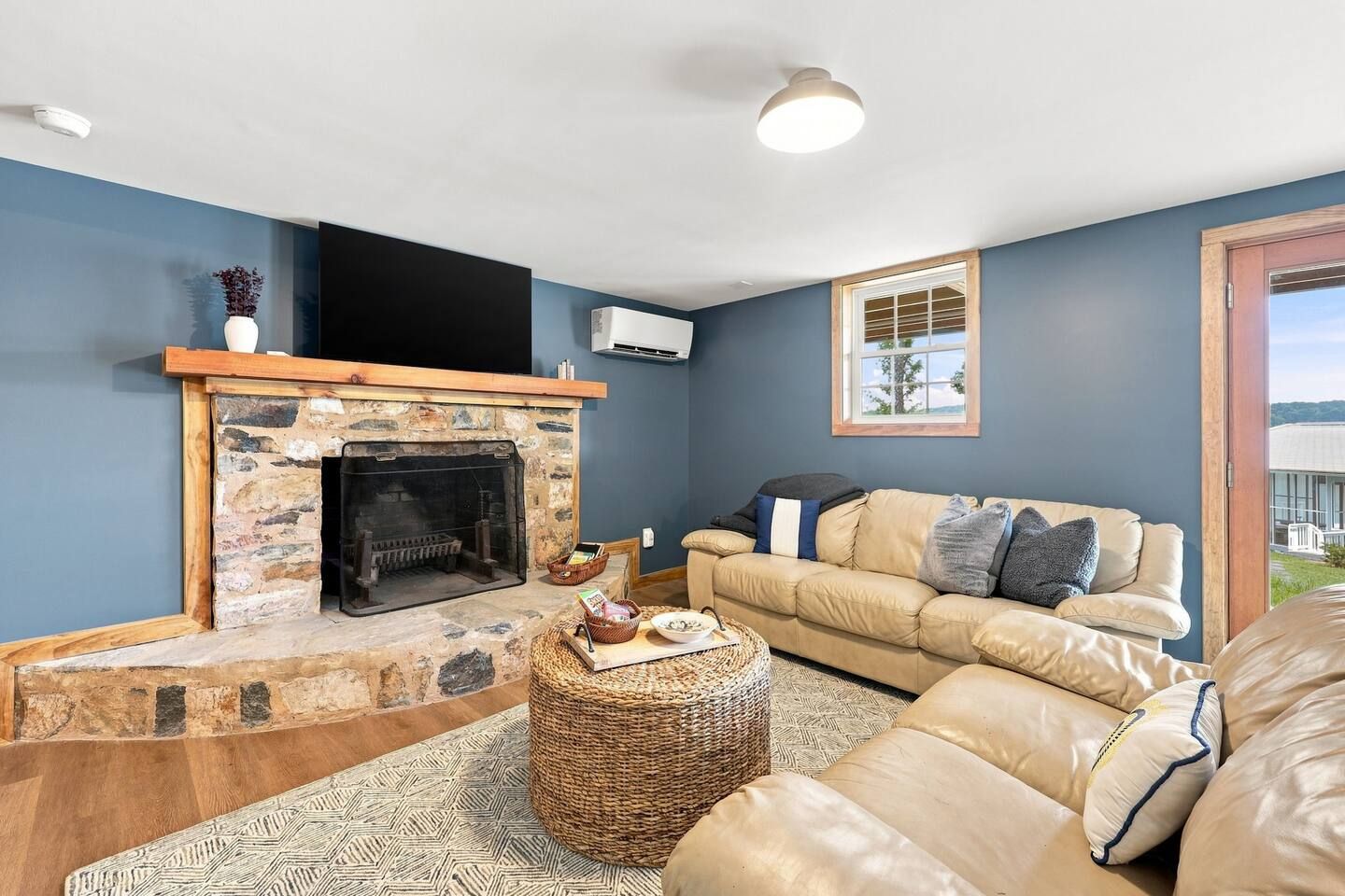 A living room with a stone fireplace, a beige sofa, and a wicker ottoman.
