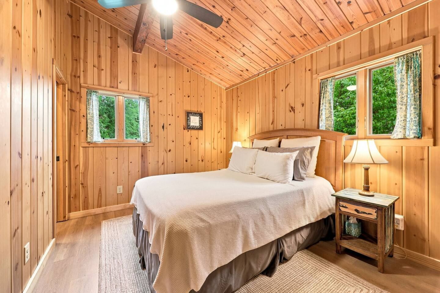 Quarto com painéis de madeira, cama, mesas de cabeceira e janelas.