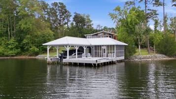 A houseboat with a covered deck, docked on a calm lake surrounded by trees.