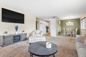 A living room with a flat-screen TV, a sofa, a dining table with chairs, and a kitchen in the background.