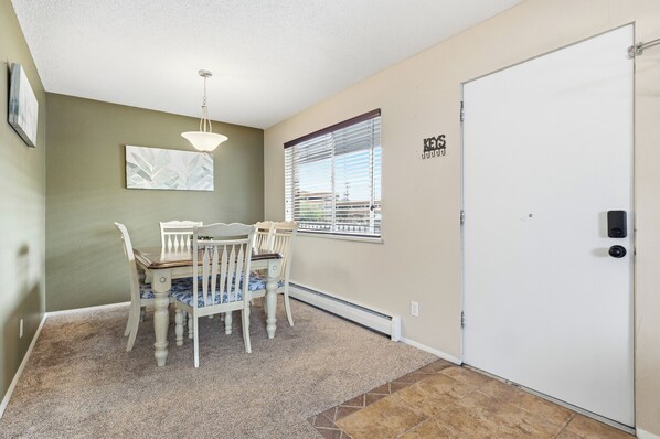 A dining area with a table and chairs, a hanging light fixture, a window with blinds, and a door with a keypad lock.