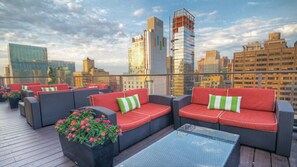 A rooftop terrace with wicker furniture and red cushions, overlooking a cityscape with modern buildings.