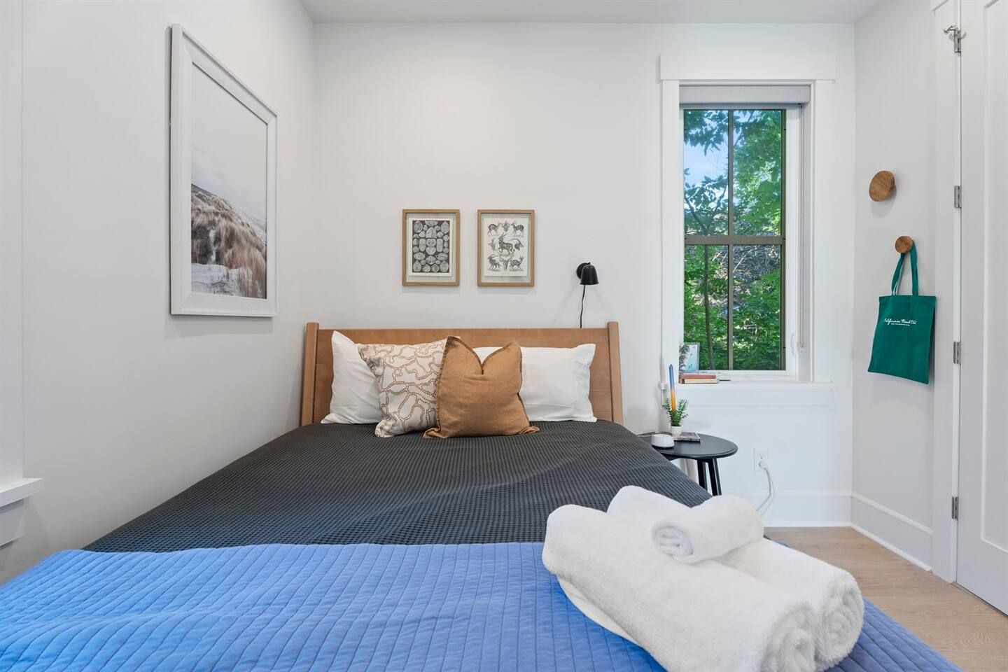 Une chambre à coucher avec un lit, des oreillers, une table de chevet avec une lampe, une fenêtre donnant sur des arbres, et un sac en tissu vert accroché au mur.