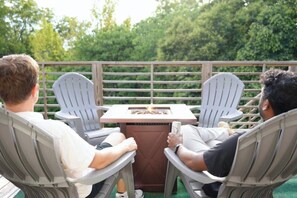 Two people seated at a table with a grill, surrounded by white chairs, on an outdoor deck with a view of trees. - Sky Deck - New Build - Downtown - Fenced - FirePit (Raleigh)