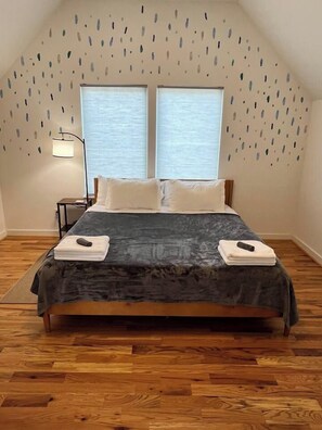 A bedroom with a large bed, two windows, a nightstand, and a wall-mounted lamp. - Sky Deck - New Build - Downtown - Fenced - FirePit (Raleigh)
