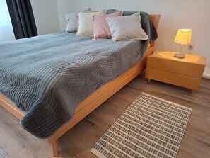 Una cama con colcha gris, una mesita de noche con lámpara y una alfombra tejida sobre piso de madera.