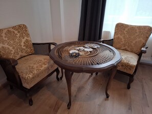Ein Zimmer mit zwei Polstersesseln mit gemustertem Stoff, einem runden Holztisch mit dekorativem Metallmittelstück und einem Fenster mit dunklem Vorhang.
