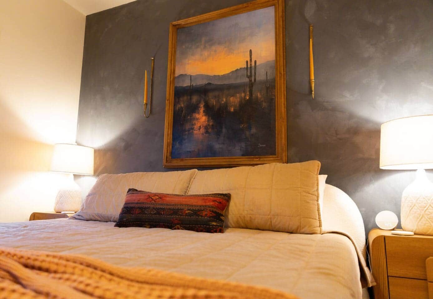 Een bed met een gekleurd kussen, een schilderij aan de muur en twee bedlampjes.