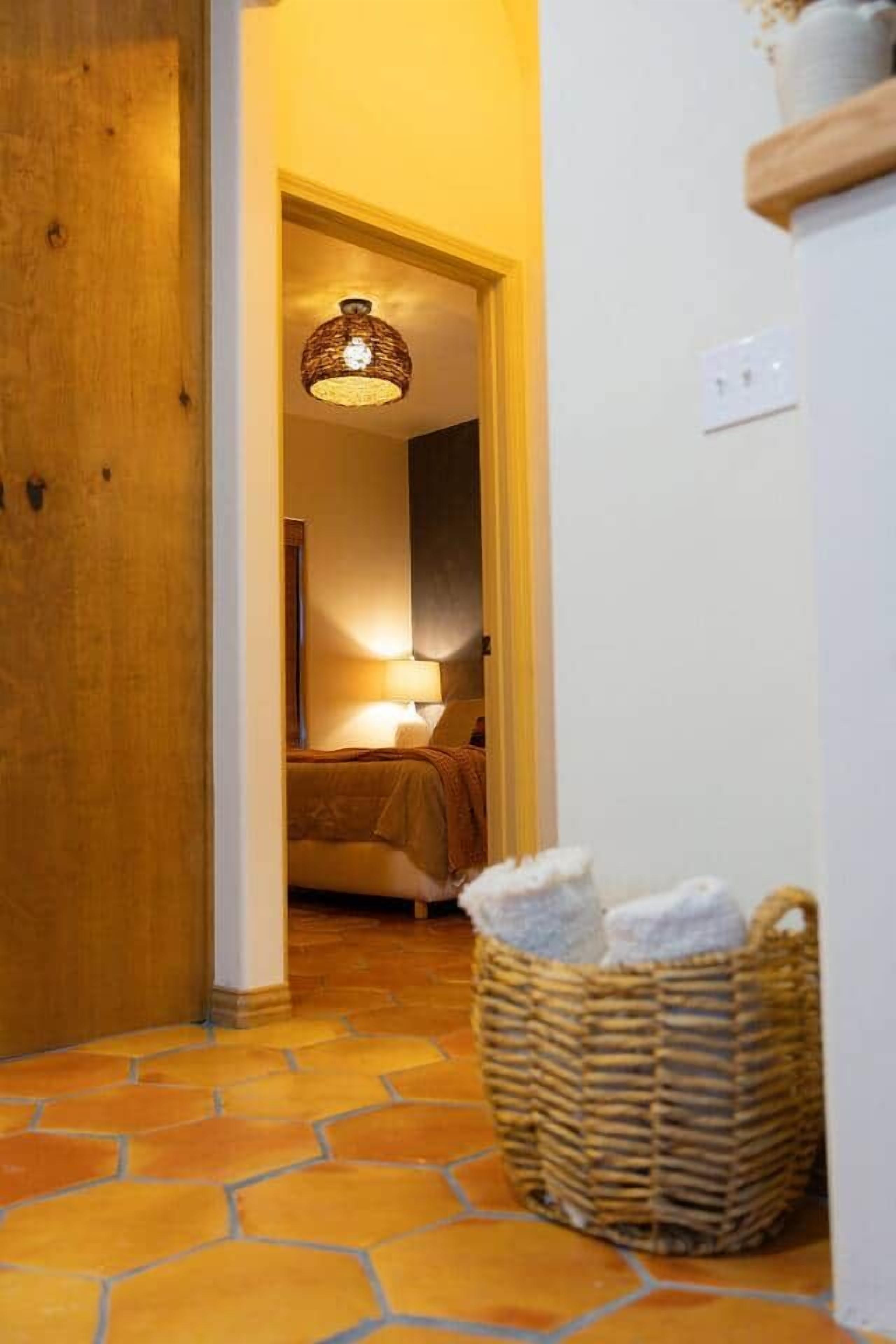 Un passadís amb una porta de fusta, una cistella de vimet i un llit amb una làmpada.