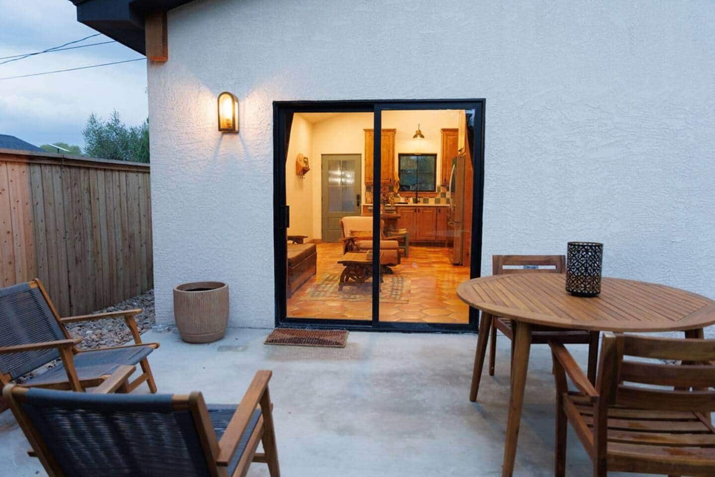 Eine Terrassenfläche mit einem runden Holztisch und Stühlen, einem Holzzaun und einer Glastür, die zu einem Innenbereich mit Küche und Sitzgelegenheit führt.