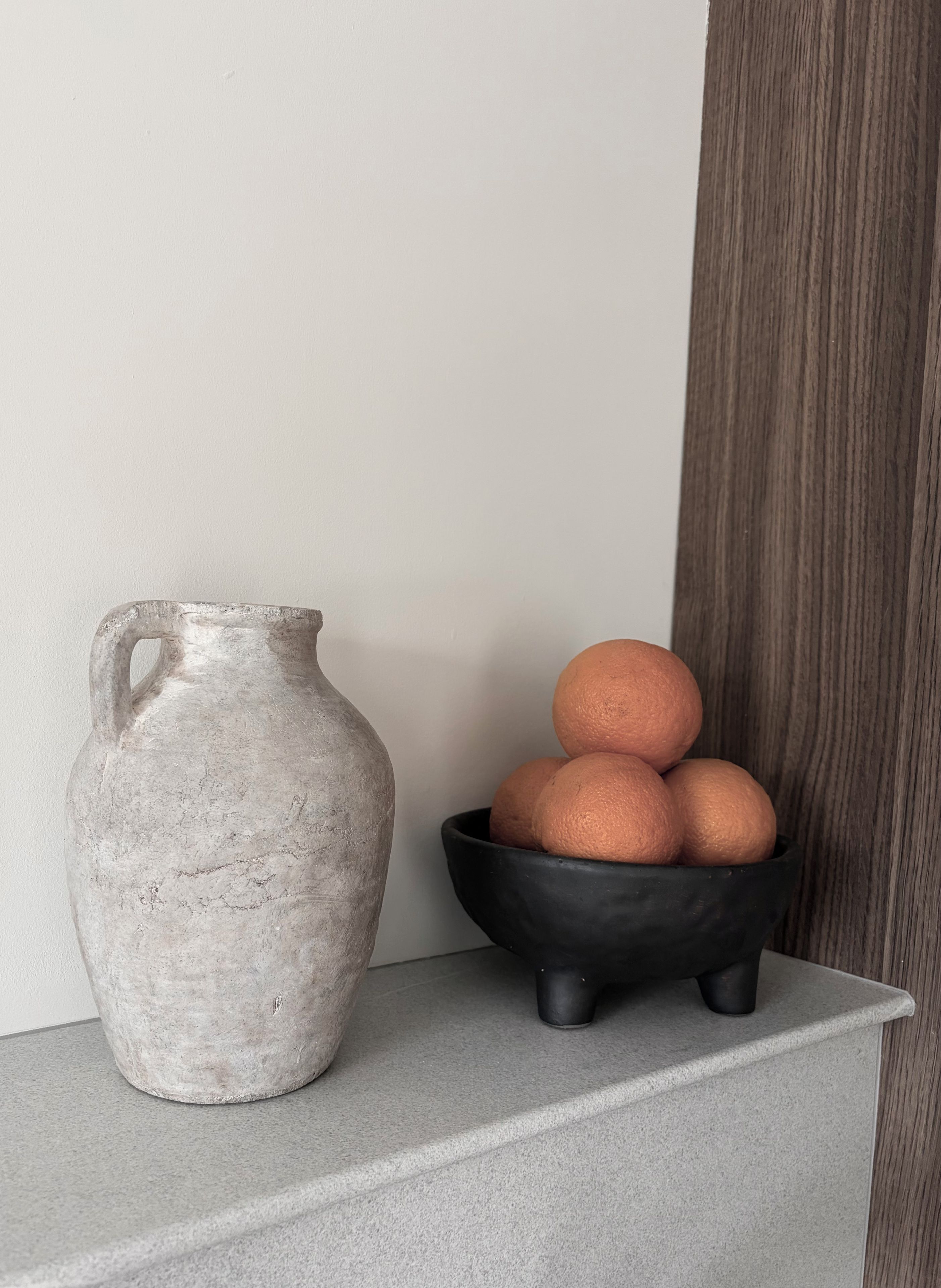Un pichet en céramique grise et un bol noir contenant des oranges, le tout posé sur un comptoir gris.