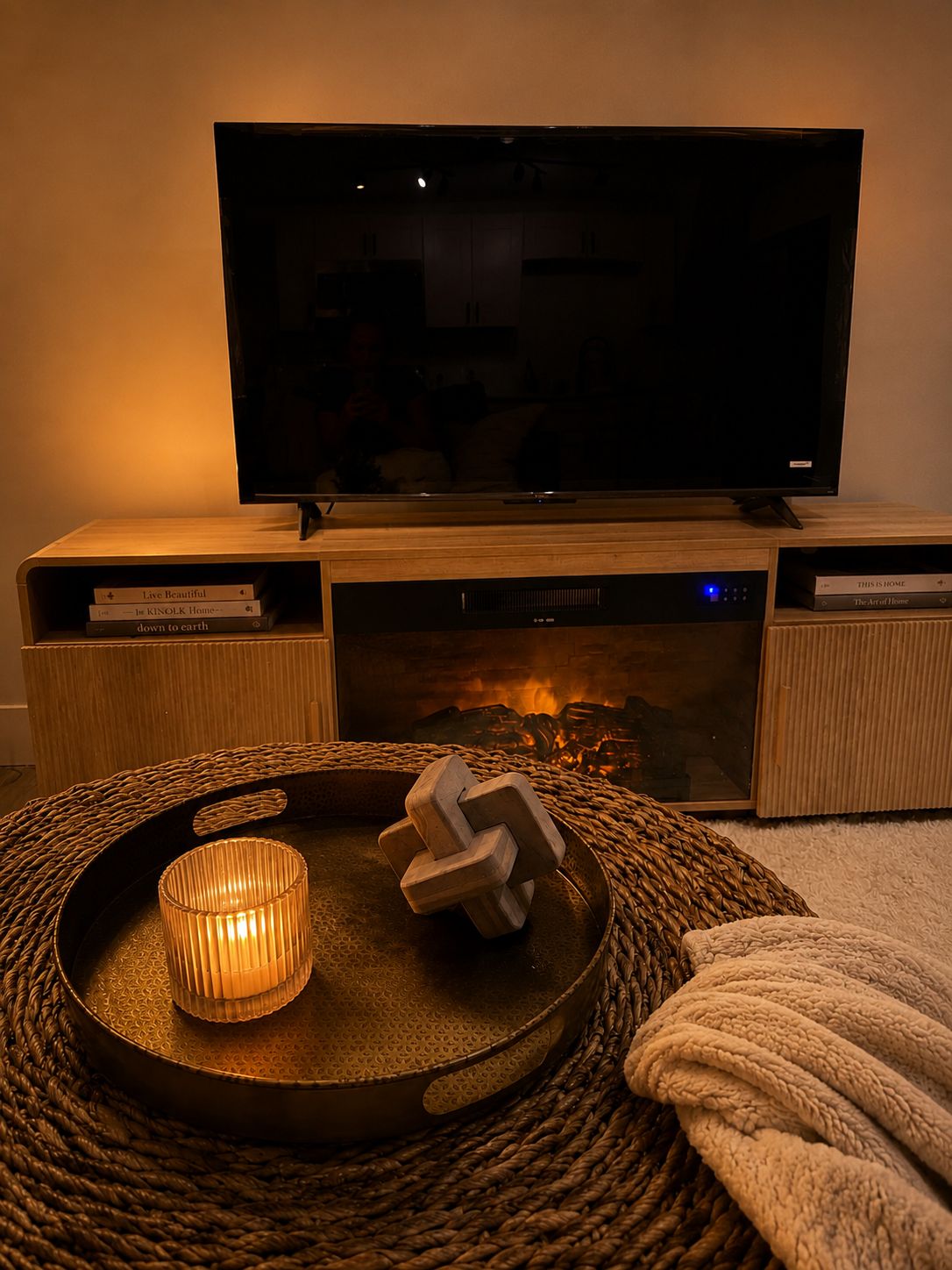 A television on a wooden stand with a fireplace below, a tray with a candle and a decorative item, and a cozy blanket on the floor.