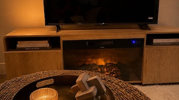 A television on a wooden stand with a fireplace below, a tray with a candle and a decorative item, and a cozy blanket on the floor.