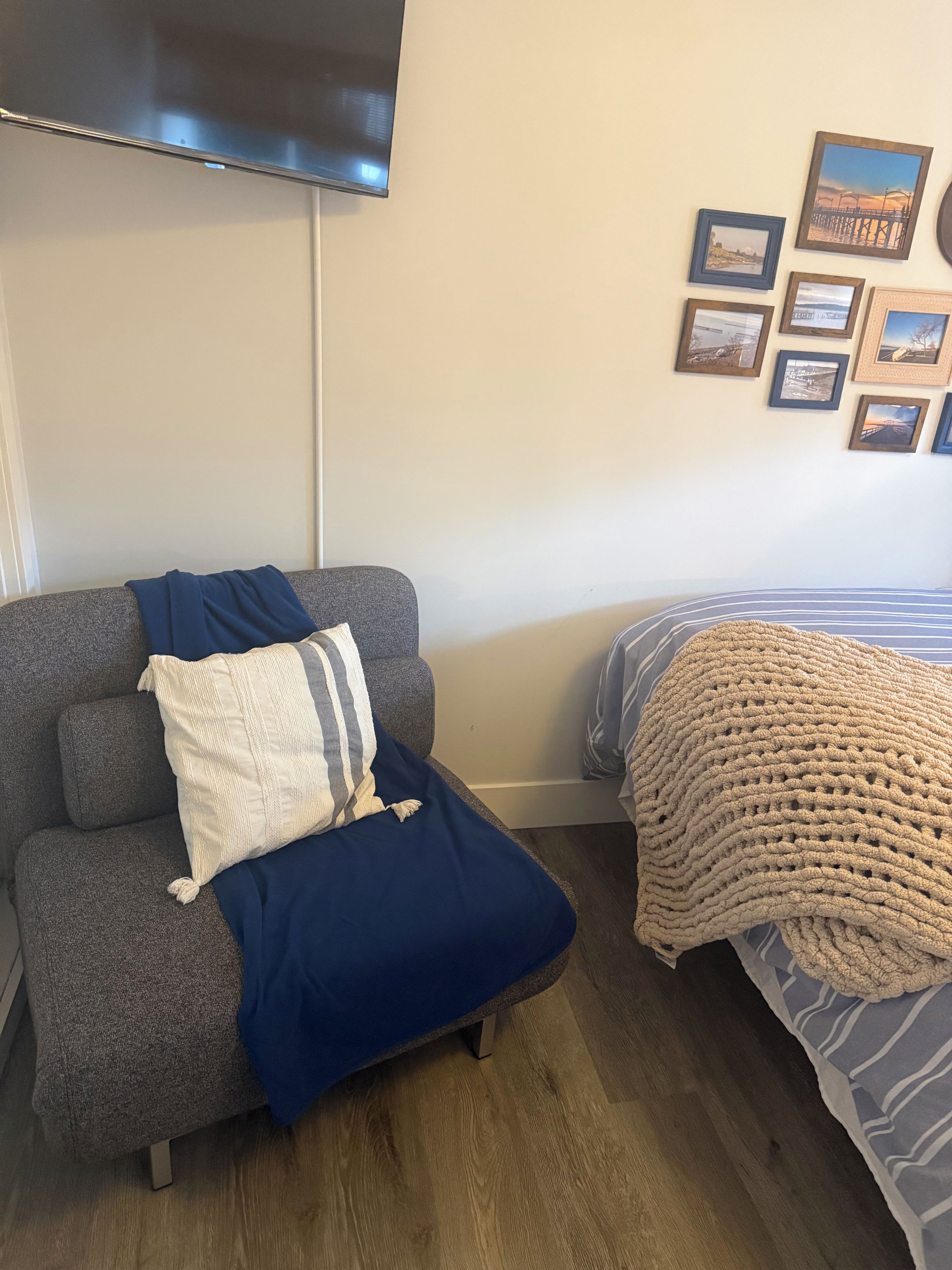 A room with a grey sofa, a blue blanket, and a bed with a striped blanket. There are framed pictures on the wall.