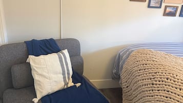 A room with a grey sofa, a blue blanket, and a bed with a striped blanket. There are framed pictures on the wall.