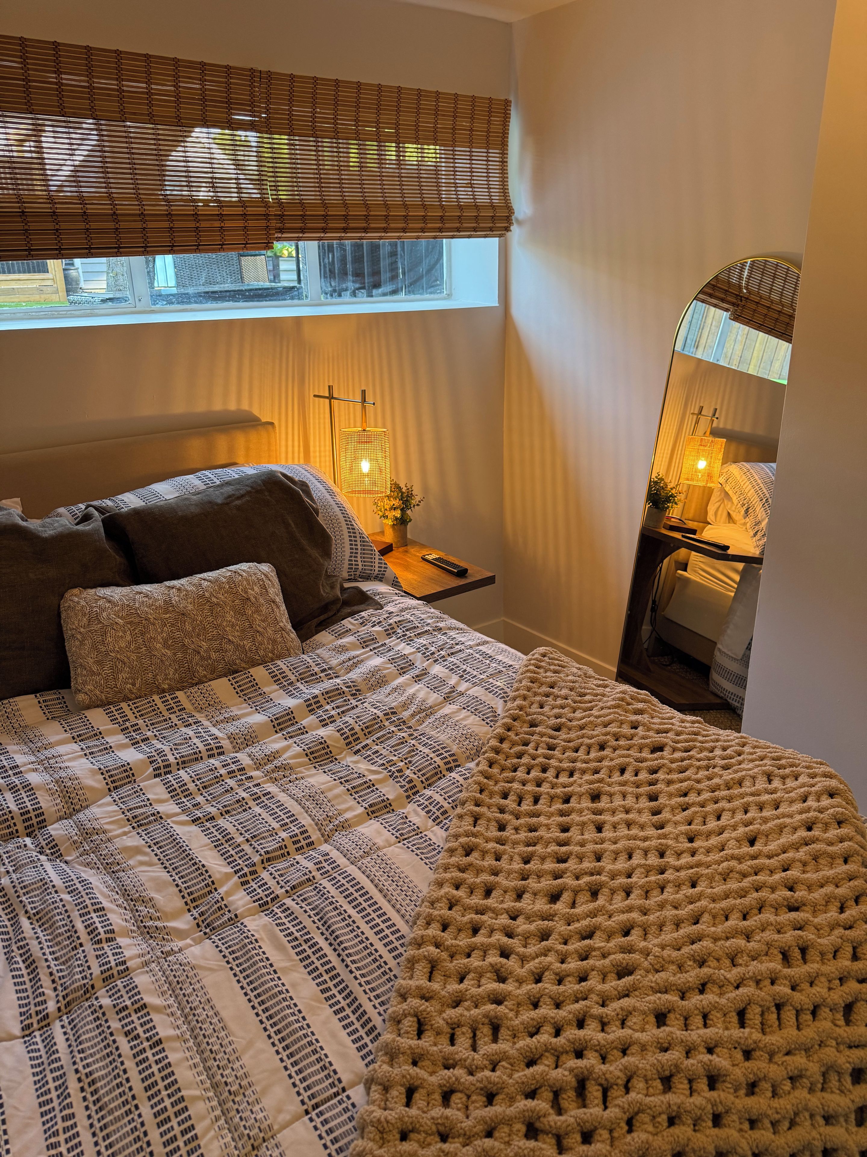 A bedroom with a bed, a bedside table with a lamp, a window with blinds, and a mirror.