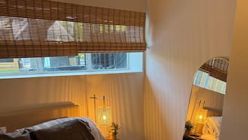 A bedroom with a bed, a bedside table with a lamp, a window with blinds, and a mirror.