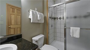 Badeværelse med toilet, håndvask og brusebad. Der er håndklæder hængende på væggen og en dør.