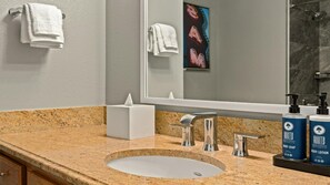 Une salle de bain avec un comptoir en granit, un lavabo, un miroir, un porte-serviettes et deux bouteilles de lotion pour le corps de la marque Roots.