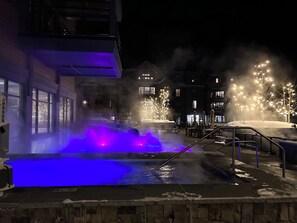 Un espace extérieur aménagé pour la nuit, avec une piscine chauffée, un sol recouvert de neige et des arbres illuminés.