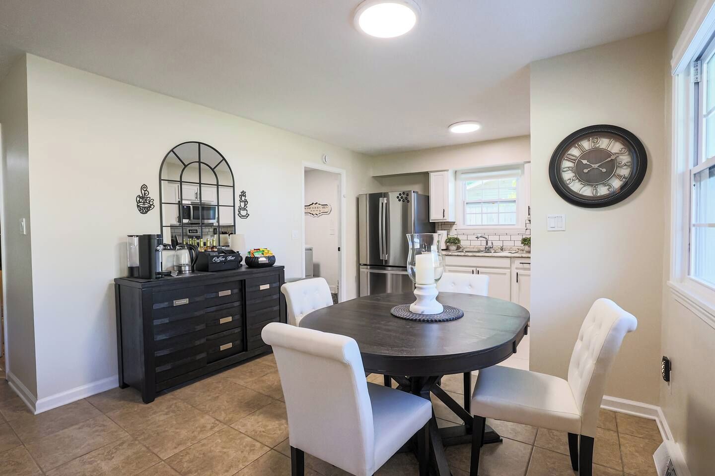 A modern kitchen with a dining area, featuring a round table, white chairs, a coffee station, and a clock on the wall.