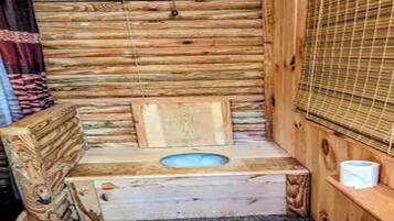 A wooden cabin interior with a built-in toilet, a window with blinds, and a shelf with a roll of toilet paper.