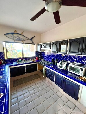 A kitchen with blue tiles, a ceiling fan, and a window with a view of palm trees. - Best Ocean View and Pool (San José del Cabo)