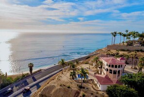 A coastal road with a view of the ocean, palm trees, and a building with a red roof. - Best Ocean View and Pool (San José del Cabo)