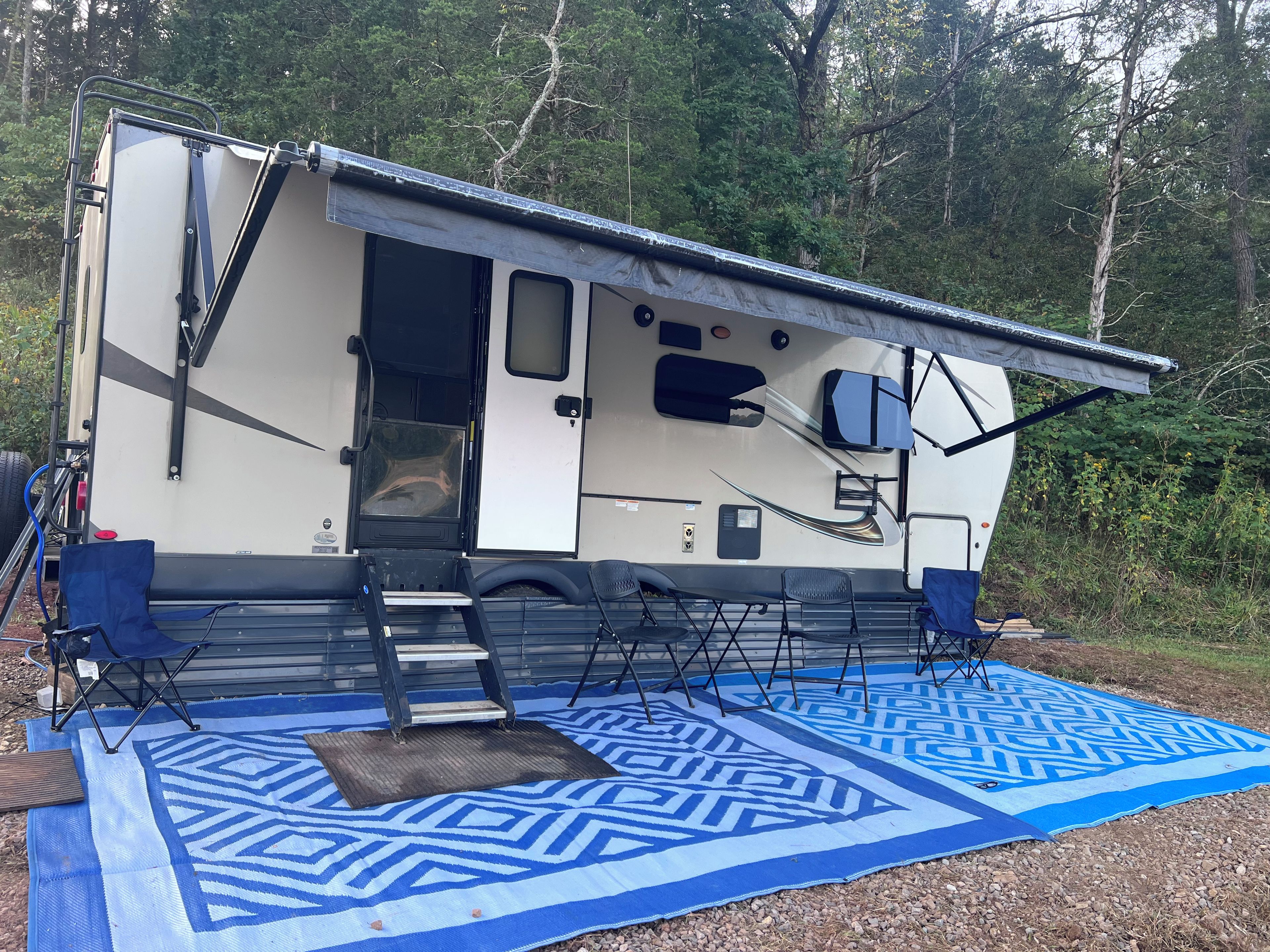 A recreational vehicle with an open door, a blue patterned mat on the ground, and a forested background.