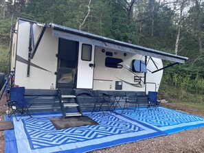 Un véhicule récréatif avec une porte ouverte, un tapis bleu à motifs posé sur le sol et un paysage boisé en arrière-plan.