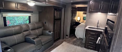 A compact RV interior with a sofa, kitchenette, and a window with a view.