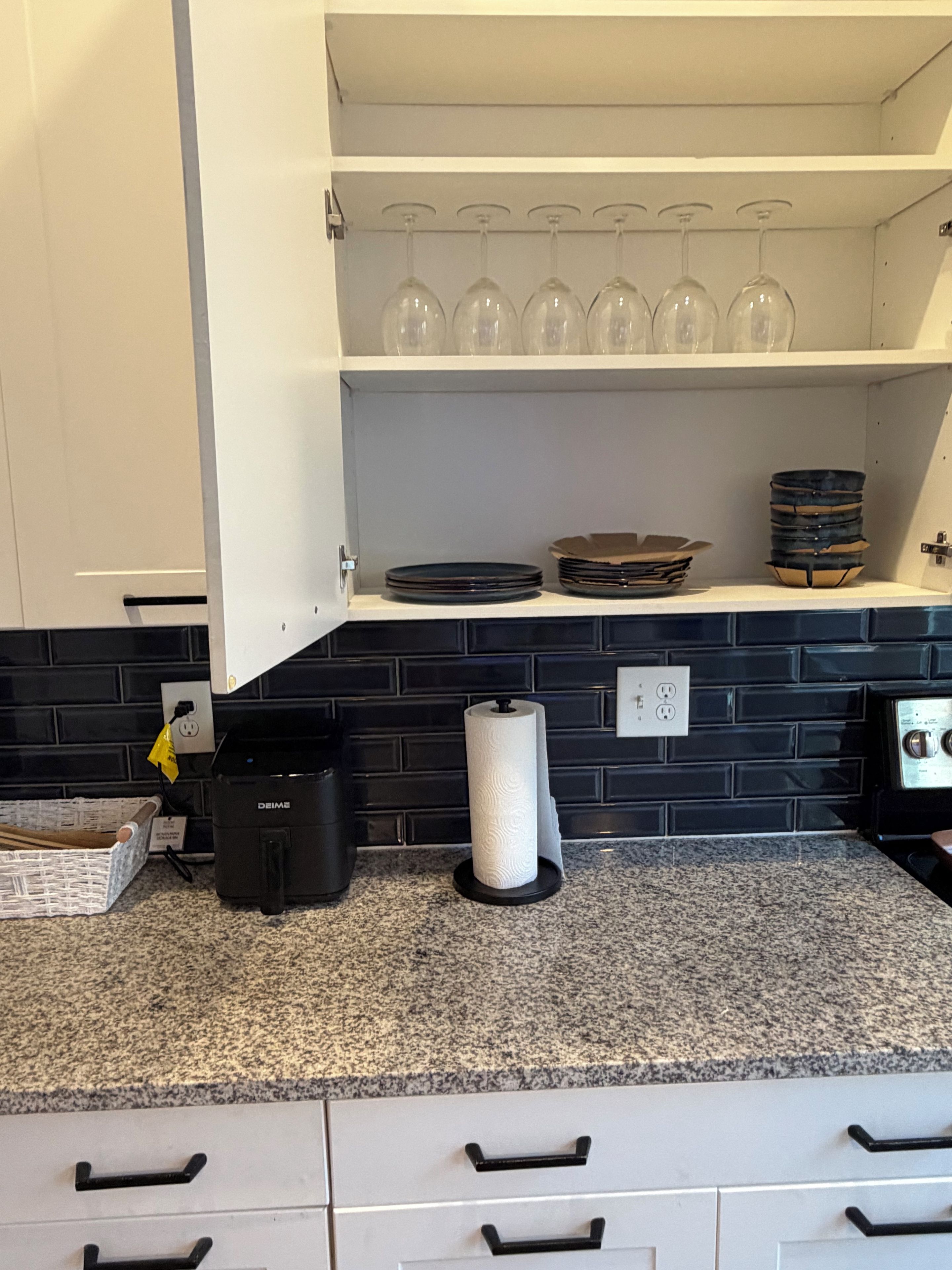 A kitchen with a black tiled backsplash, a countertop with a paper towel dispenser, a coffee maker, and a set of wine glasses on a shelf.