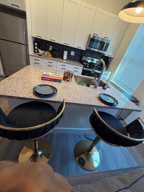 A kitchen with a granite countertop, stainless steel appliances, and two black bar stools. - The Midnight Escape/Fast&Furious Suite (Atlanta)