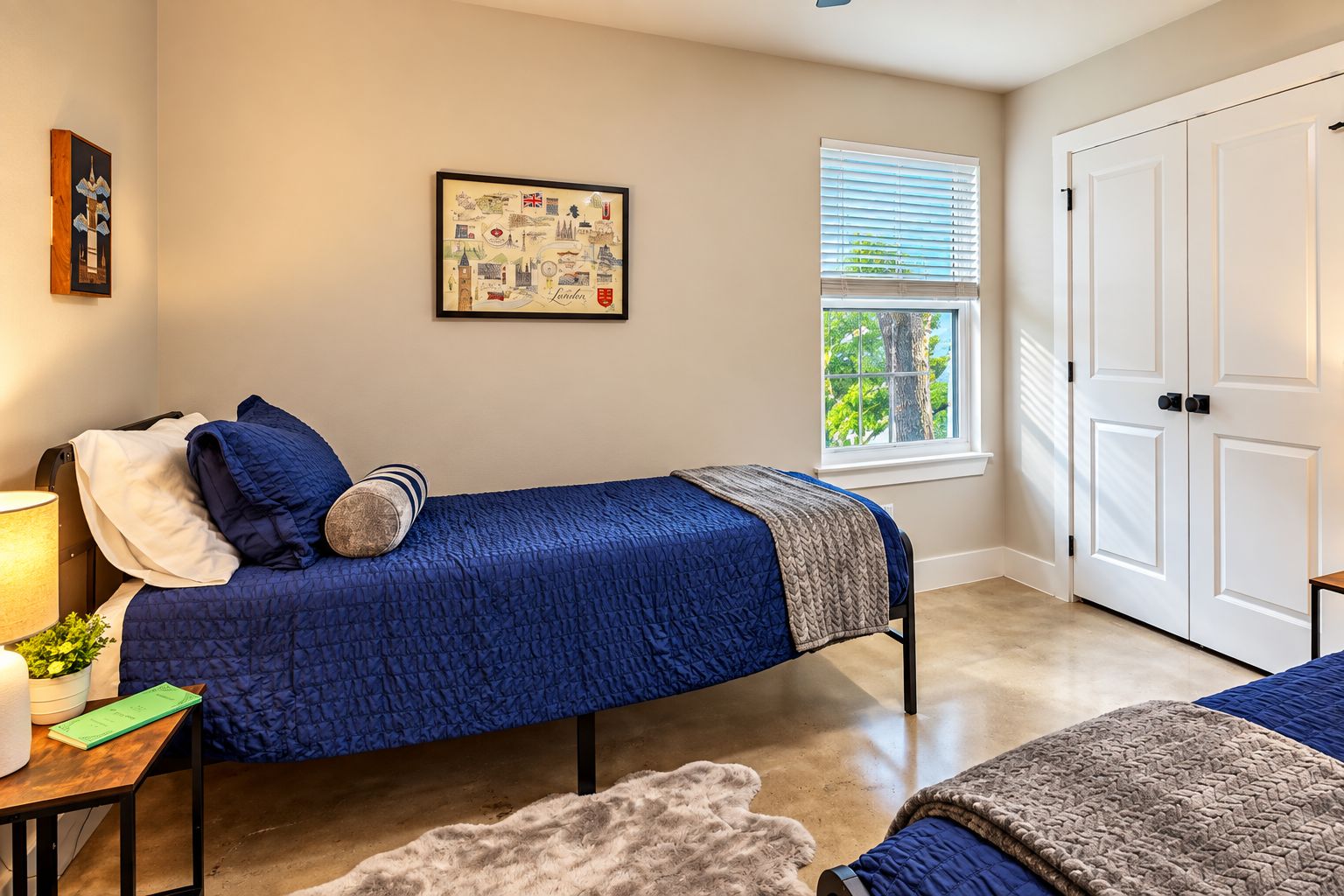 A bedroom with a bed, a bedside table with a lamp, a framed picture on the wall, and a window with blinds.