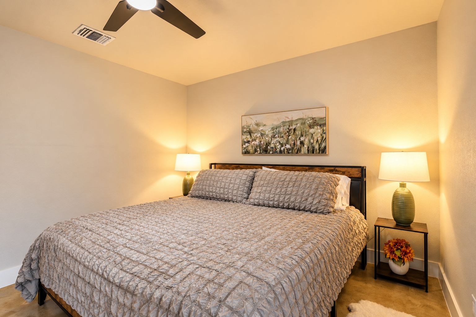A bedroom with a bed, two bedside lamps, a ceiling fan, and a wall-mounted painting.