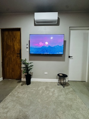 A room with a mounted TV displaying a mountain scene, a potted plant, a small stool, and an air conditioning unit. - Premiun 1-Bed Apart in Pinilla - Tamarindo - Avellanas w AC / Pool / Rancho BBQ (Pinilla)