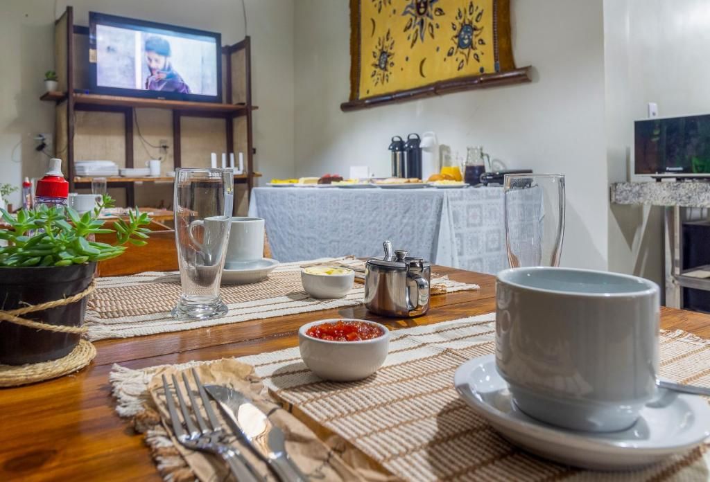 Uma área de jantar com uma mesa posta para dois, uma televisão ao fundo e uma parede com um quadro decorativo.