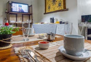 A dining area with a table set for two, a television in the background, and a wall with a decorative painting. - Guest House 4 Anjinhos (Porto Seguro)