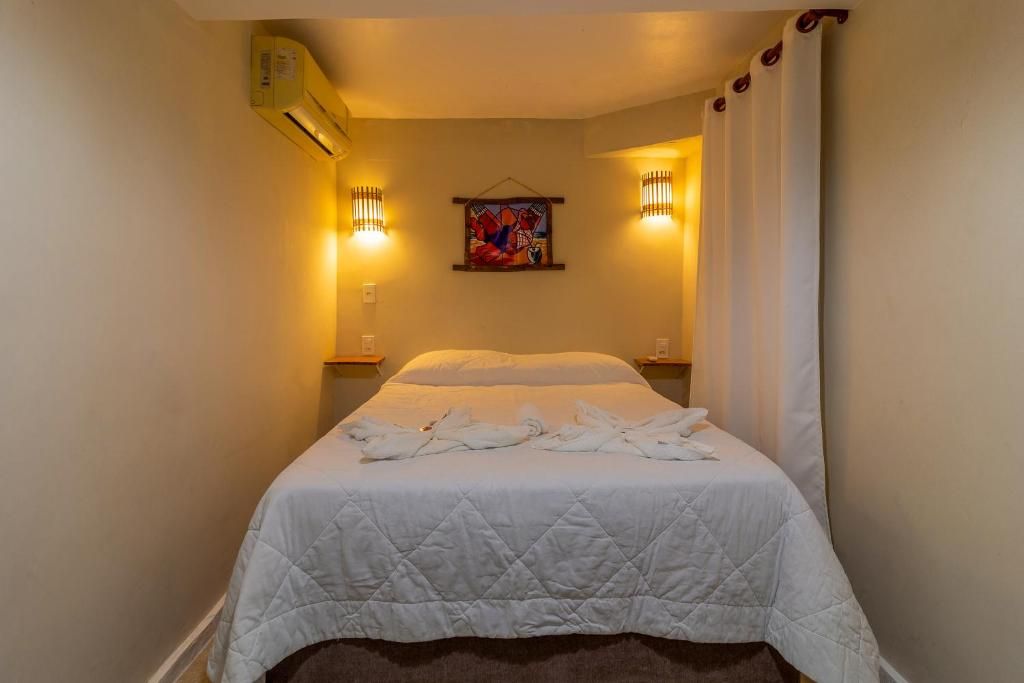 Quarto de hotel com cama, ar-condicionado fixado na parede, duas arandelas e um quadro.