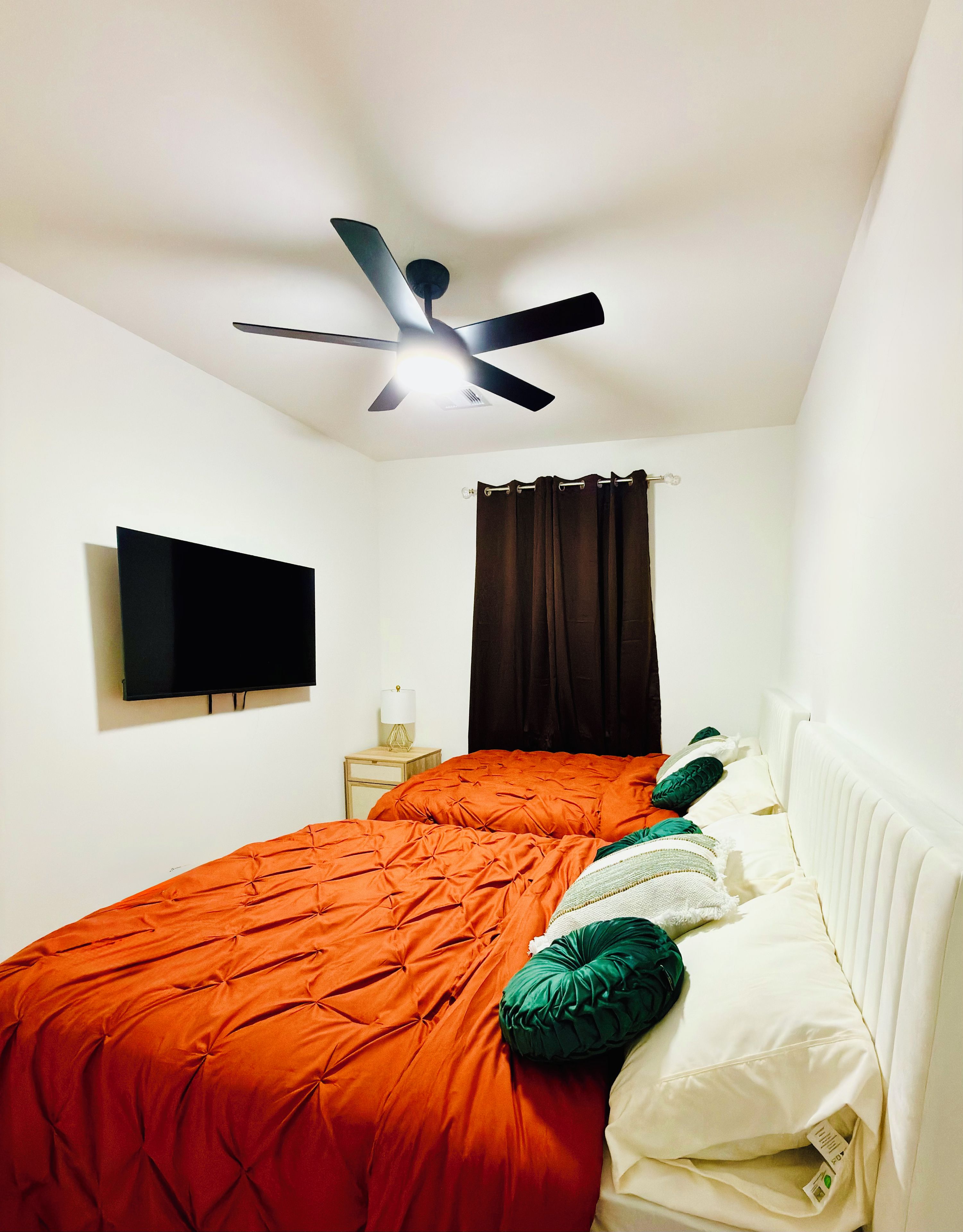 A bedroom with a bed, a ceiling fan, a TV, and a window with curtains.