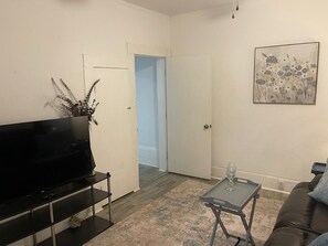 A living room with a flat-screen TV on a black stand, a coffee table with a glass, and a brown leather sofa. - Charming Cozy Midtown Cottage near Lake Ella/FSU/FAMU/CIVIC CENTER/TMH (Tallahassee)