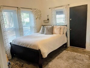 A bedroom with a bed, curtains, a door, and a wall-mounted light fixture. - Charming Cozy Midtown Cottage near Lake Ella/FSU/FAMU/CIVIC CENTER/TMH (Tallahassee)